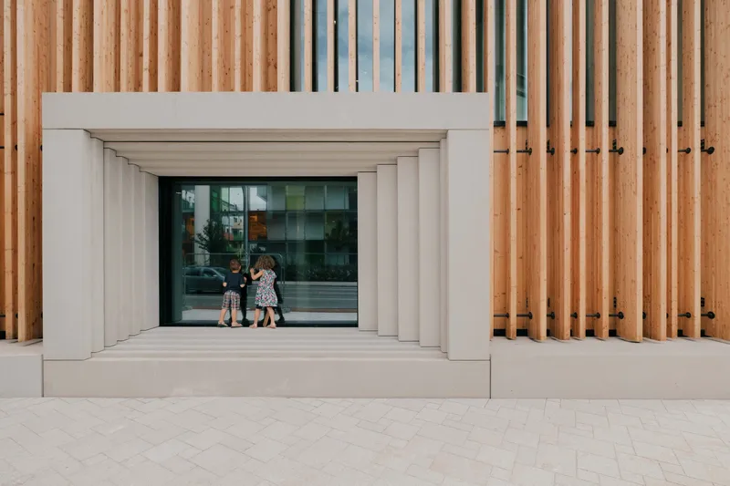 Facade detail with vertical timber slats and white stone portal framing the glazed entrance as children enter