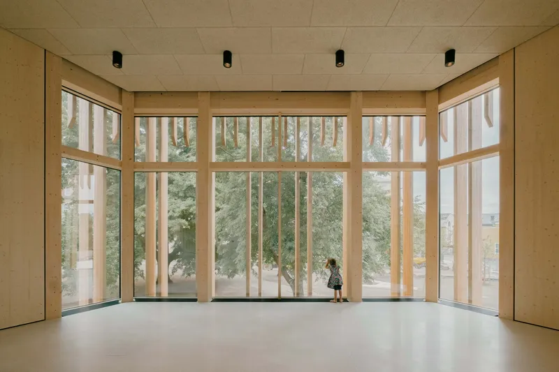 Sunlit interior room with floor-to-ceiling windows framing trees outside and a child silhouetted against the glass