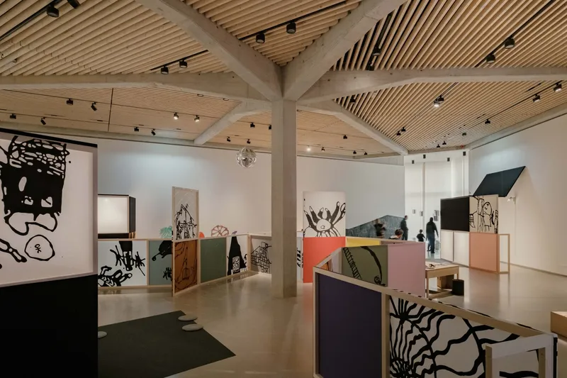 Gallery interior with timber slat ceiling, concrete columns, and exhibition partitions displaying artworks