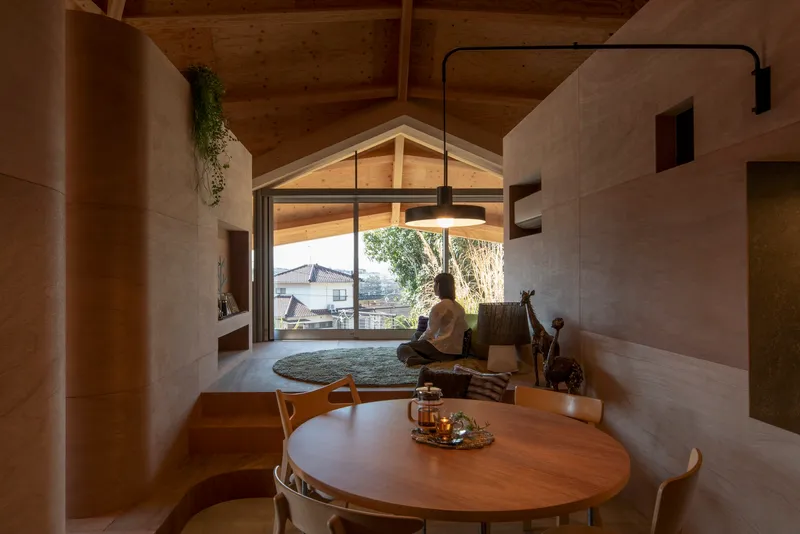 Living area with plywood partitions and round dining table where a person sits on a rug near the gabled window