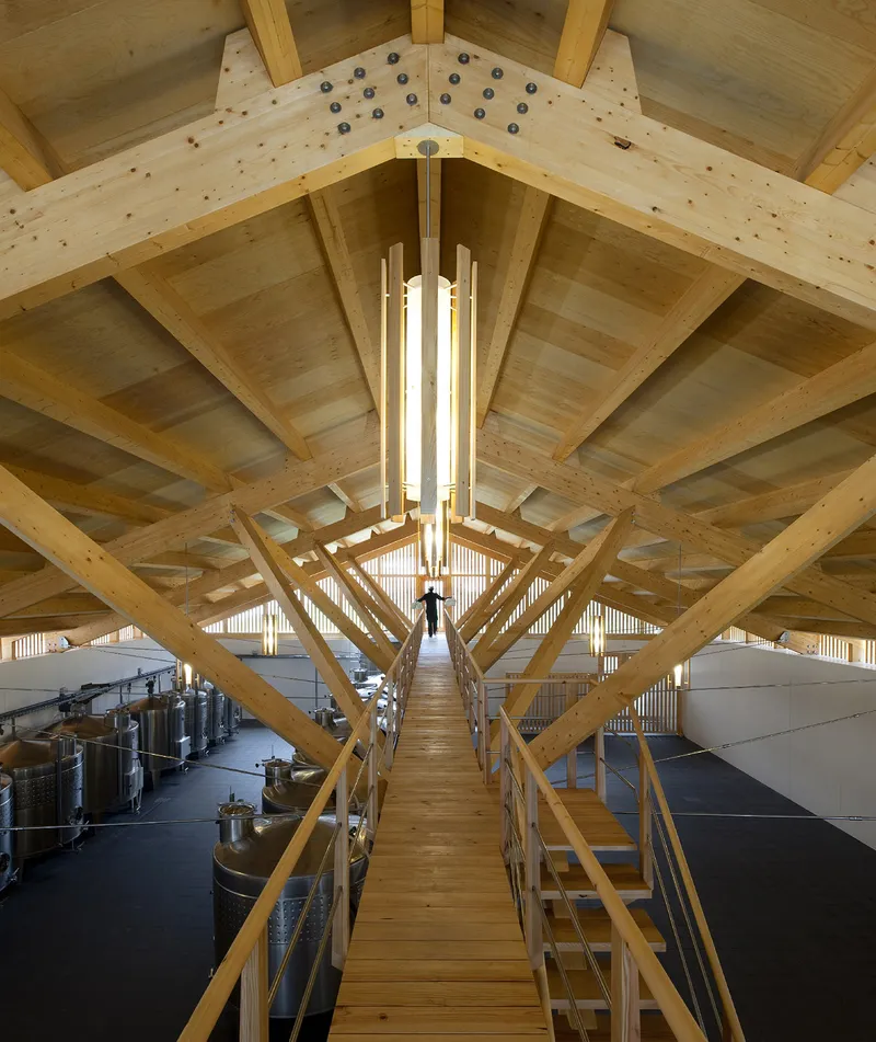 Suspended timber walkway beneath exposed wood truss roof with person descending between production tanks