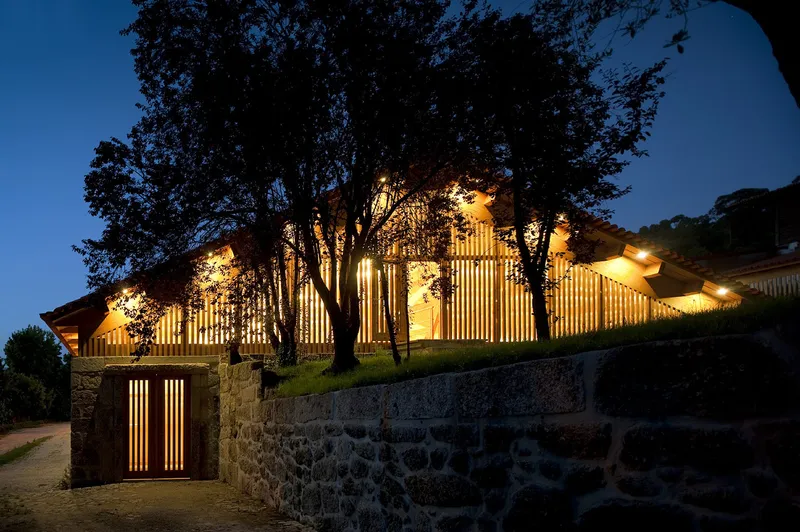 Night view of the glowing timber screen facade behind stone retaining walls and silhouetted trees
