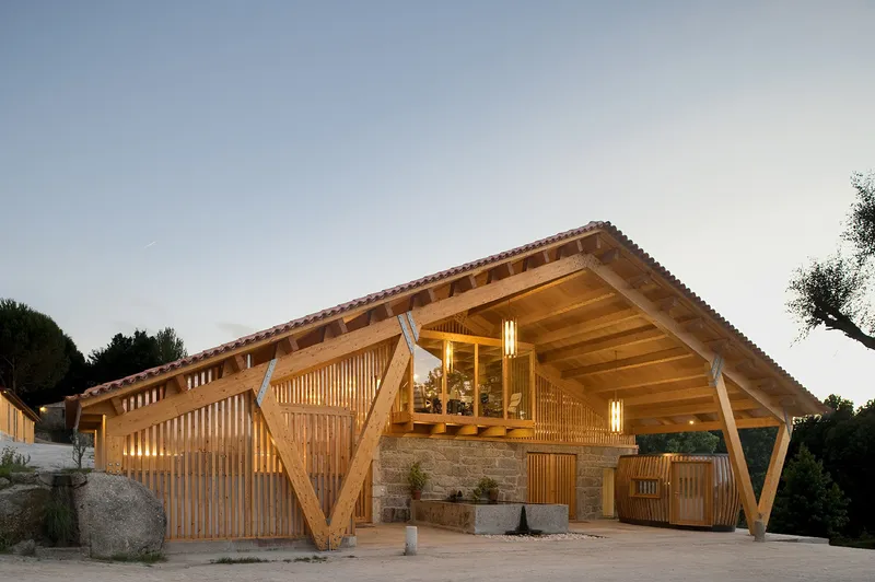 Entrance facade with V-shaped timber columns and vertical wood slats framing the glazed upper level at twilight