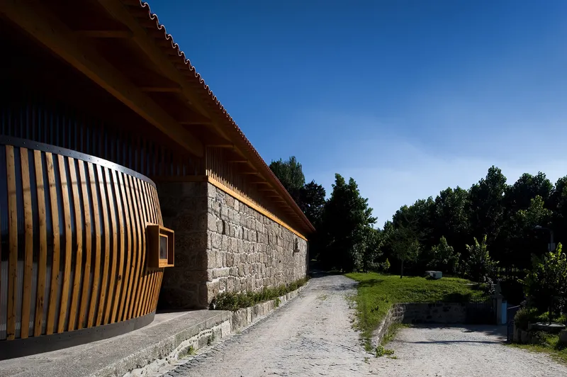 Curved timber screen wrapping the stone masonry wall along a gravel path under bright afternoon sun