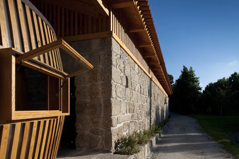 Long facade with granite stone base wall and timber slatted upper volume beneath deep eaves at dusk