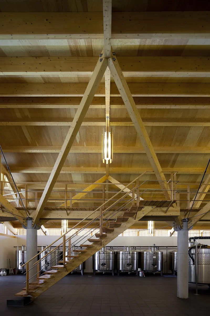 Interior view of exposed timber A-frame trusses and beams above an open staircase and stainless fermentation tanks