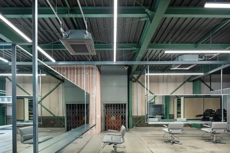Open salon floor with green steel beams, ribbed wood partitions, and rows of salon chairs under linear lighting