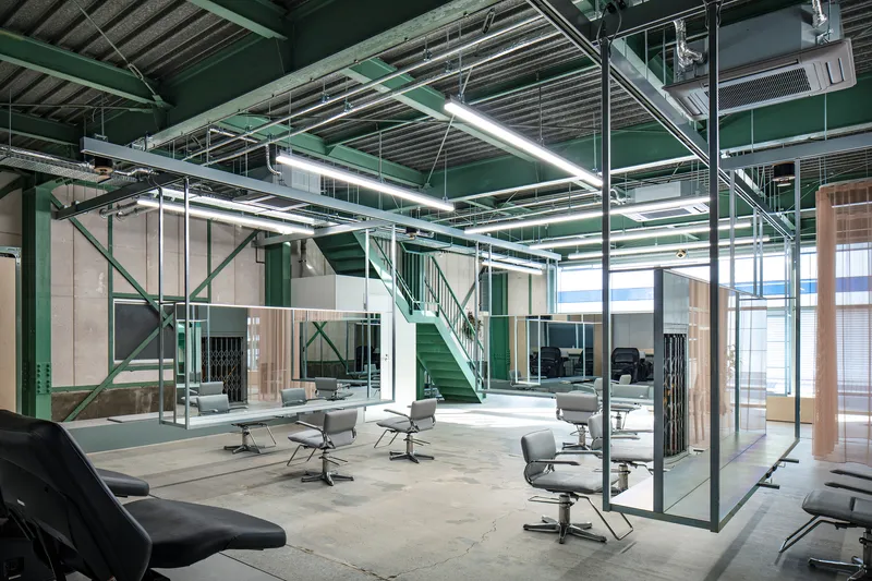 Double-height salon interior with green steel mezzanine, glass partitions, and rows of salon chairs under fluorescent tubes