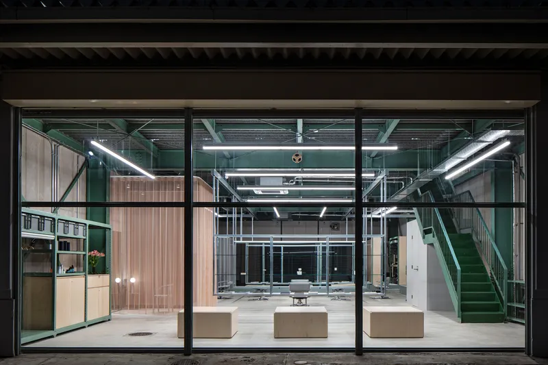Night view through full-height glass facade showing green steel staircase and exposed mechanical ductwork