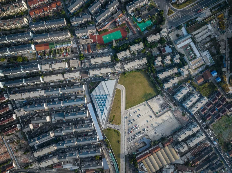 Drone view showing the angular roof geometry and adjacent parking lot within the dense urban fabric