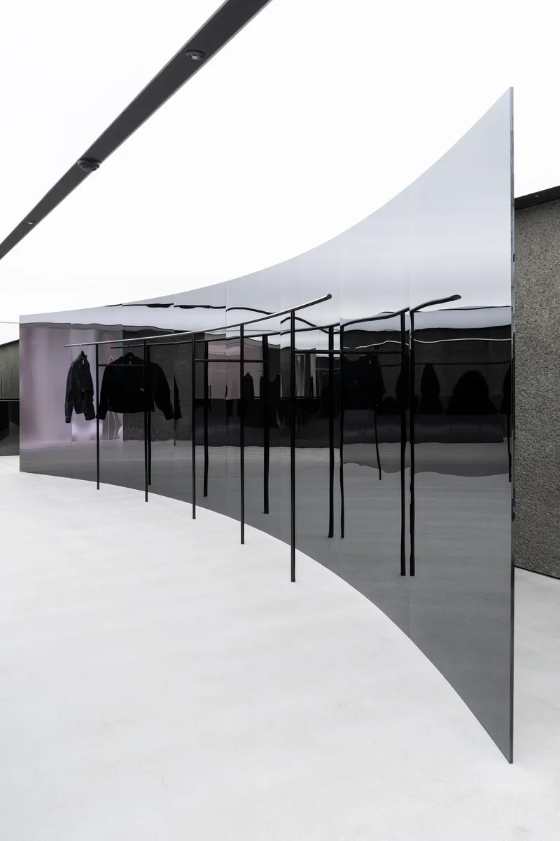 Angled view of clothing display racks behind dark glass with textured stone wall under white fabric ceiling