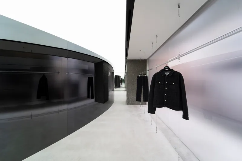 Display hallway with curved dark glass wall opposite illuminated white shelving and hanging denim jacket