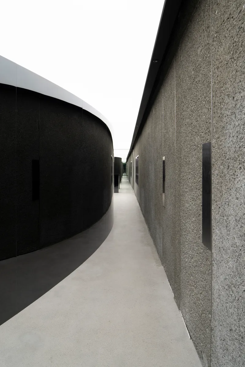 Narrow corridor between curved black wall and textured stone surface with vertical slot openings