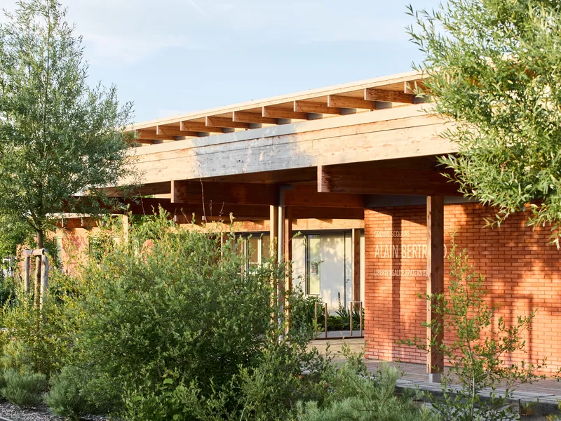 Entrance canopy with exposed timber beams and red brick walls framed by young olive trees