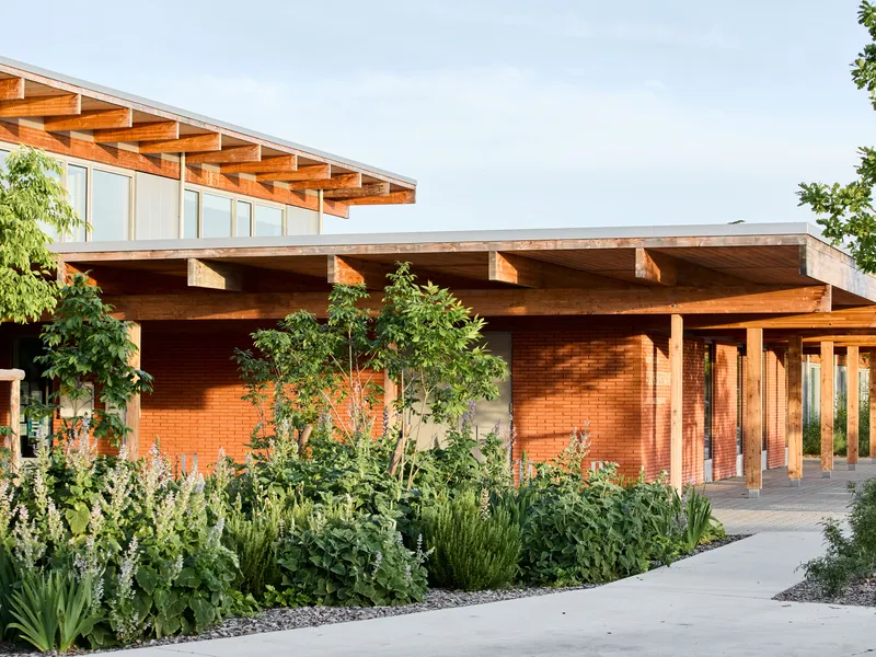 Exterior facade showing exposed timber structure above red brick base surrounded by planted beds