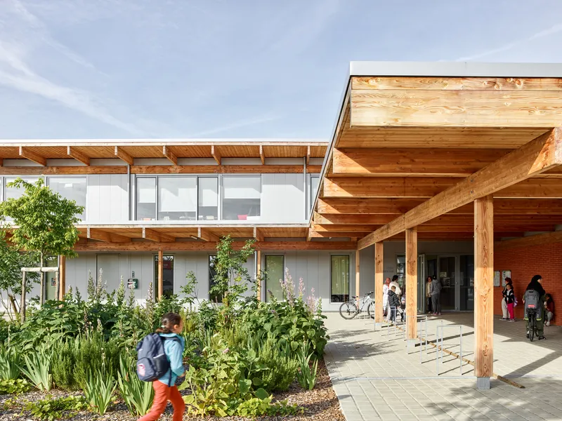 Covered entry walkway with exposed timber beams and columns beside a planted garden bed with people arriving