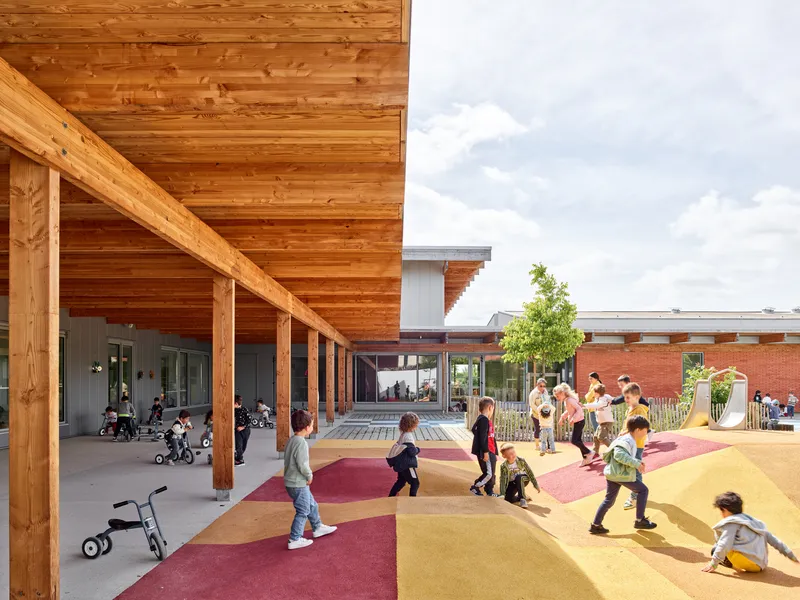Covered timber colonnade sheltering children playing on colorful rubber surfacing adjacent to a brick volume