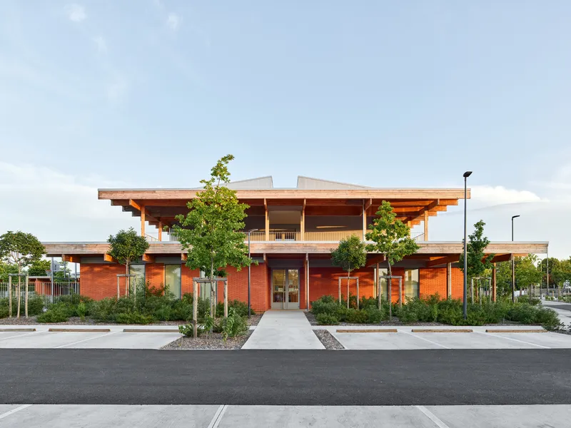Street-facing elevation with red brick walls and exposed timber roof structure framed by young trees