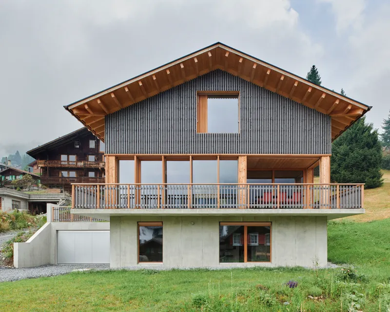 Gabled facade with vertical metal cladding and exposed timber eaves above a concrete base and grass slope