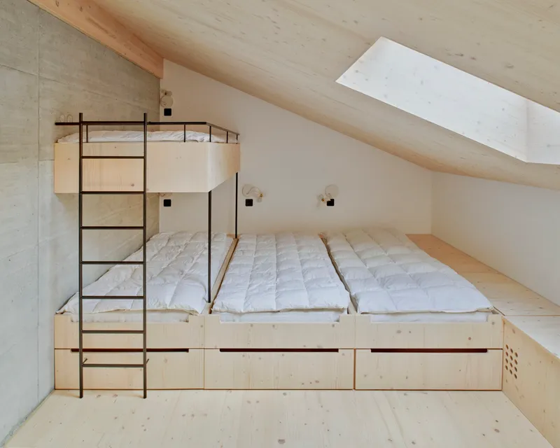 Sleeping loft with built-in timber platform bed and metal ladder beneath sloped ceiling with skylight