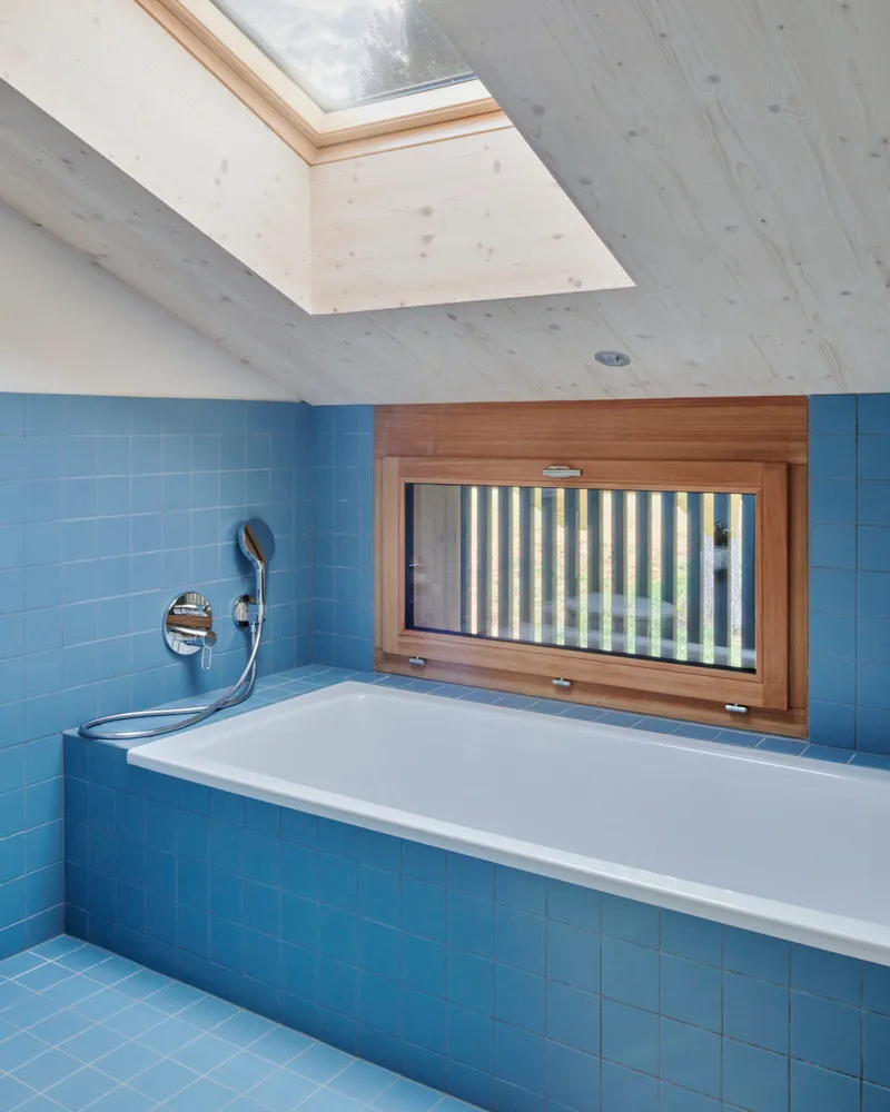 Bathroom with blue tile surround, white bathtub, timber-framed window and skylight above exposed wood ceiling