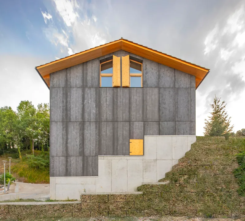Gable end facade with grey concrete panels, timber framed window and exposed hillside behind