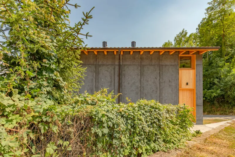 Concrete wall with timber rafter overhangs and timber door viewed through overgrown hedgerow in summer
