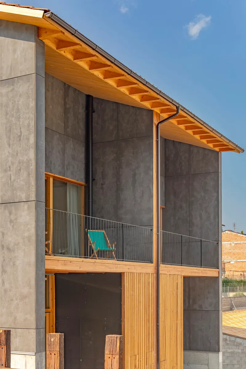 Facade detail showing concrete panels with timber window frames and exposed rafter eaves under blue sky