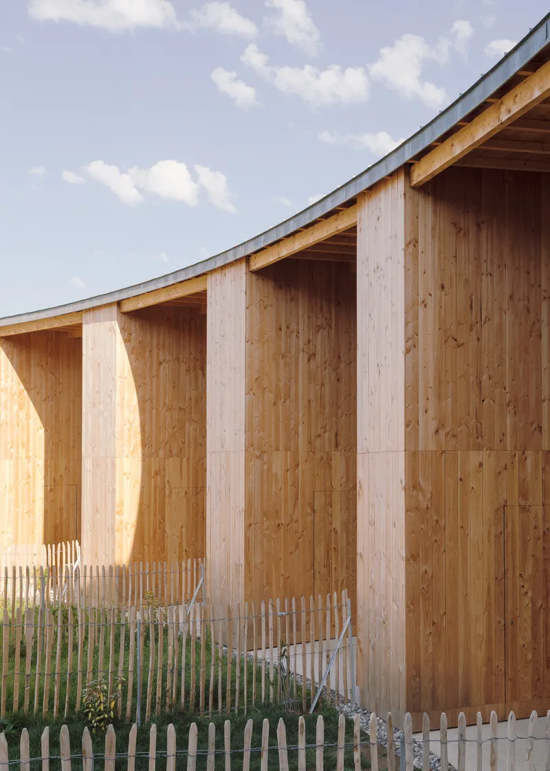 Close-up of vertical timber cladding with chamfered pilasters and white picket fence under a curving metal roof