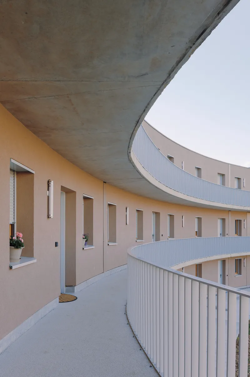 Curved exterior corridor with concrete soffit, cream stucco walls, and vertical slit windows at dusk