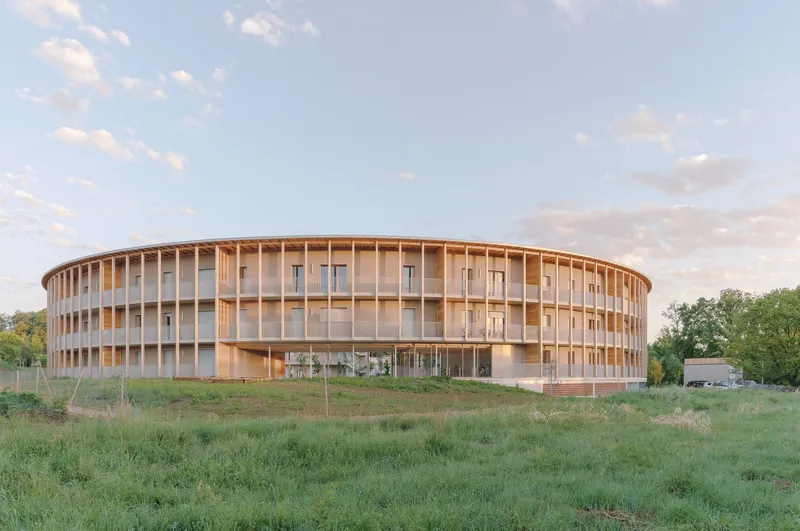 Circular timber-framed building with vertical louvres and glass enclosure at dusk in an open field