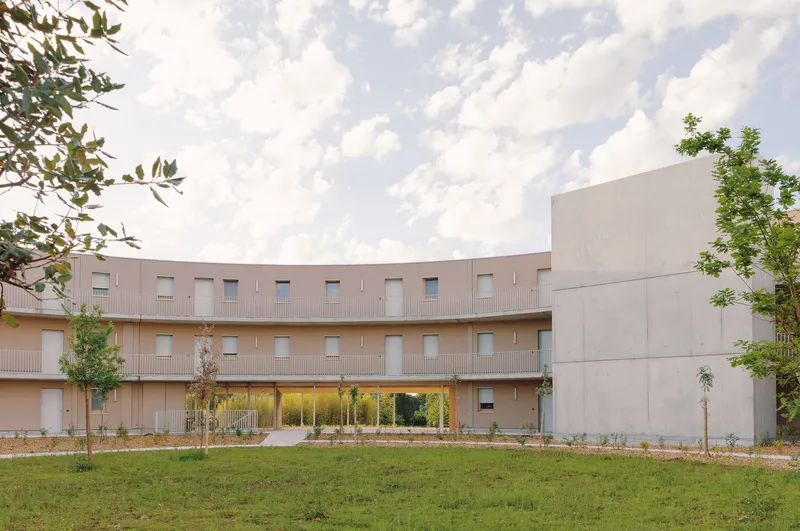 Curved timber facade with recessed entrance and sapling trees under a cloudy summer sky