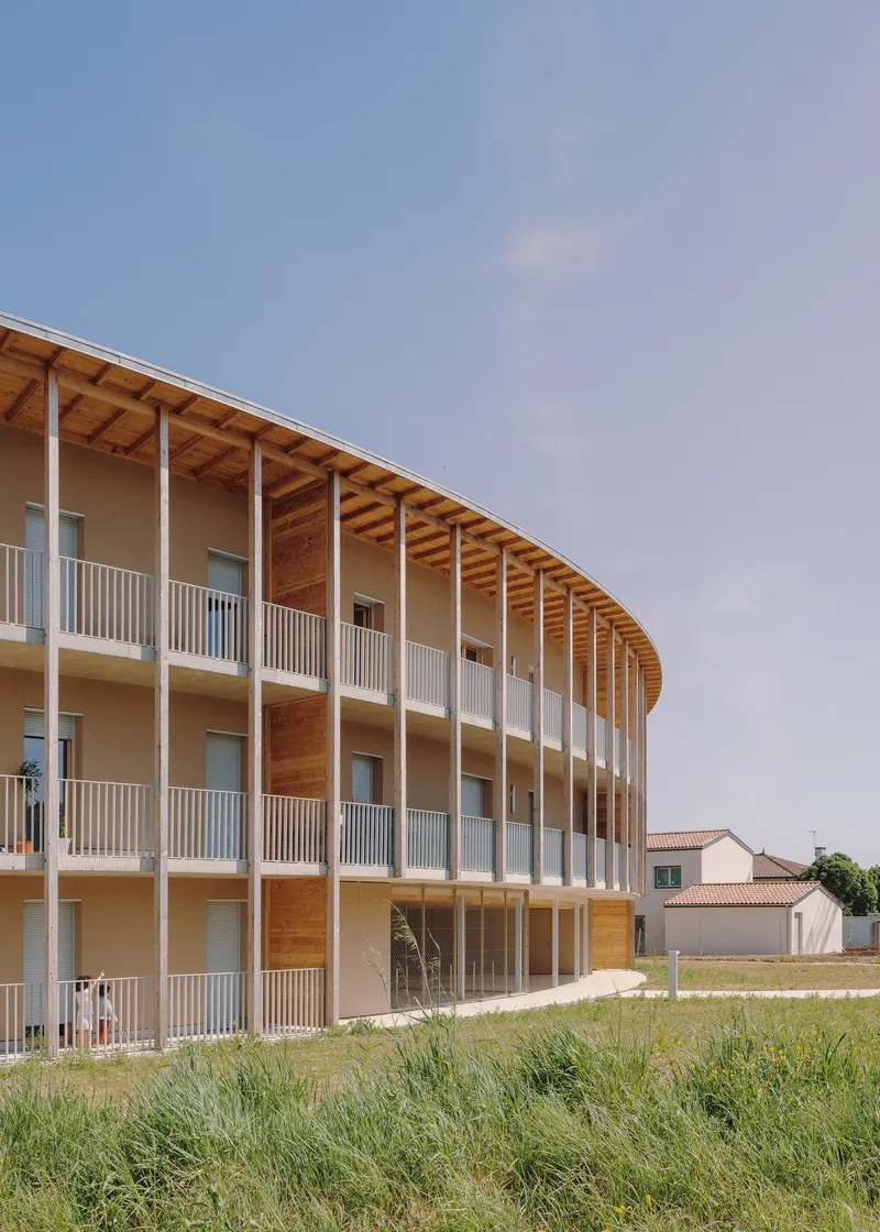 Curved timber facade with three levels of white metal balconies and exposed rafter ends under clear sky