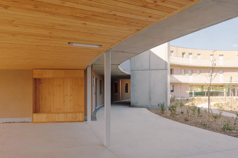 Ground floor walkway with timber ceiling, concrete beam and curved concrete wall framing the courtyard beyond