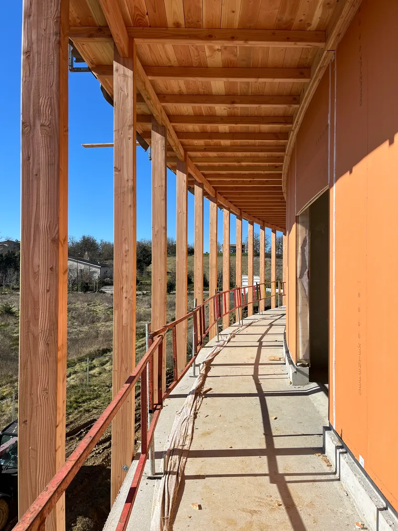 Exterior walkway with exposed timber beams, red metal railing and concrete floor casting afternoon shadows