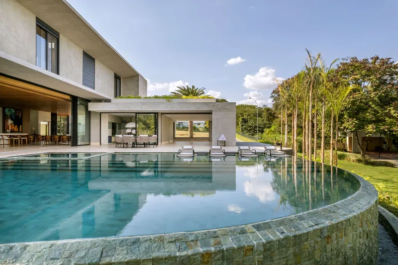 Curved pool with stone tile edge reflecting sky and clouds alongside open-plan living pavilion