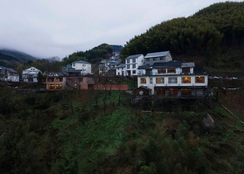 White painted timber structure with illuminated windows nestled among traditional houses on a forested hillside at dusk