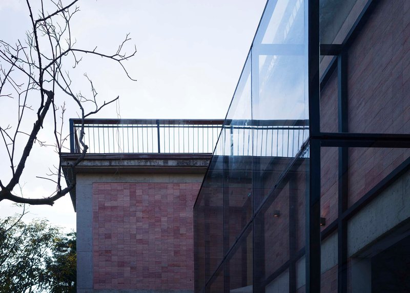 Glass facade reflecting rammed earth tile wall and bare tree branches under an overcast sky
