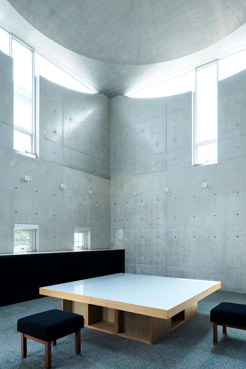 Vaulted concrete space with clerestory windows illuminating walls and low table with stools below