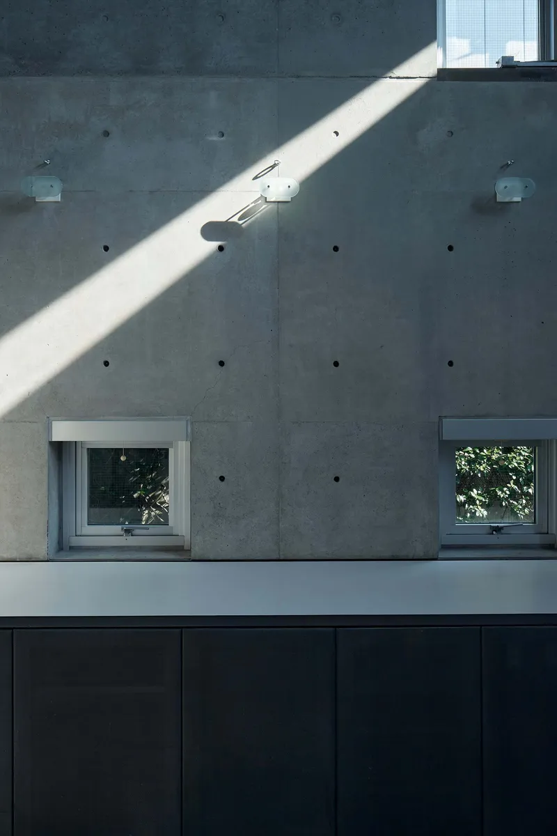 Board-formed concrete wall with two square windows and a diagonal shaft of afternoon sunlight above dark cabinetry