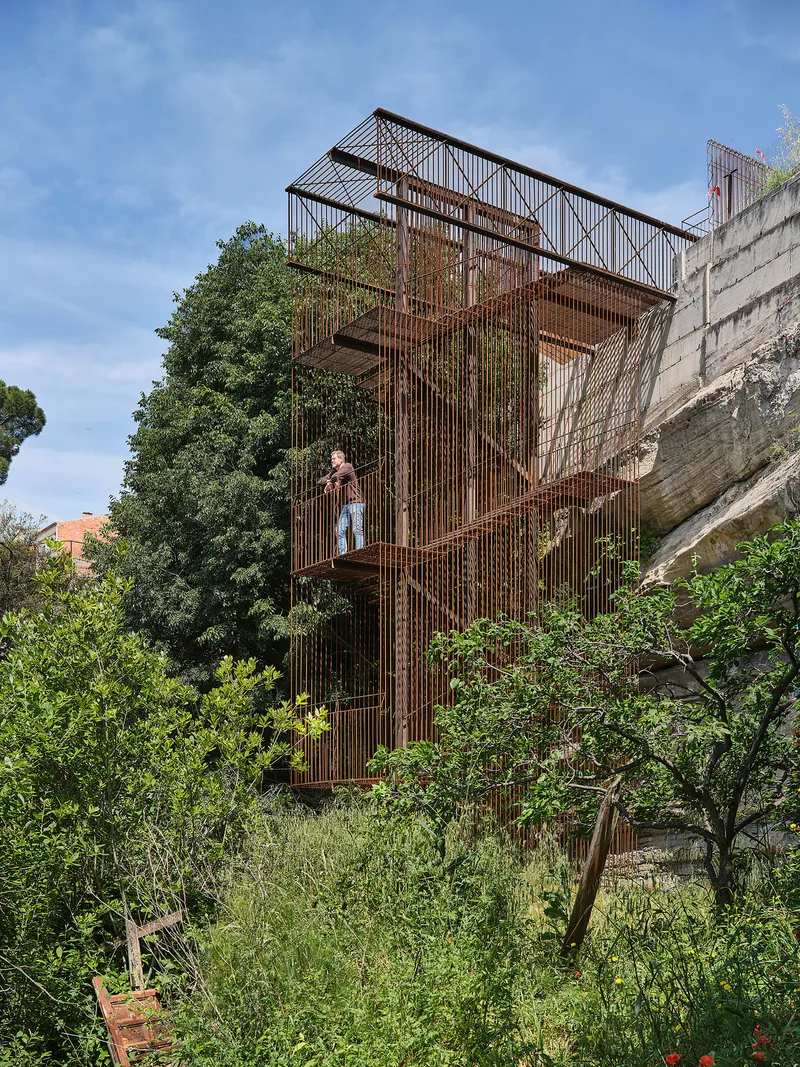 Vertical corten steel staircase tower with a person standing on a platform surrounded by dense vegetation