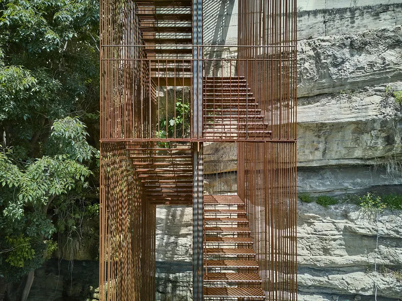 Vertical metal screen staircase with slatted treads against a stratified stone wall in dappled sunlight