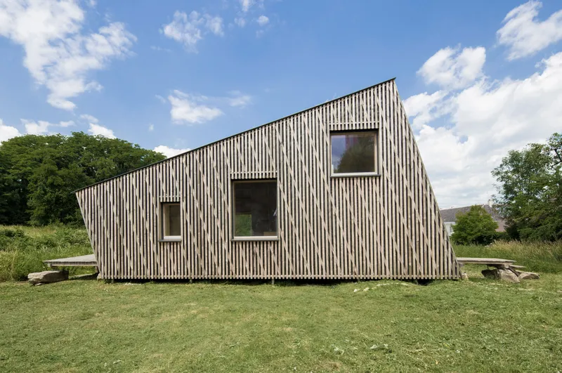 Gabled volume clad in vertical timber slats with scattered window openings in a grassy clearing
