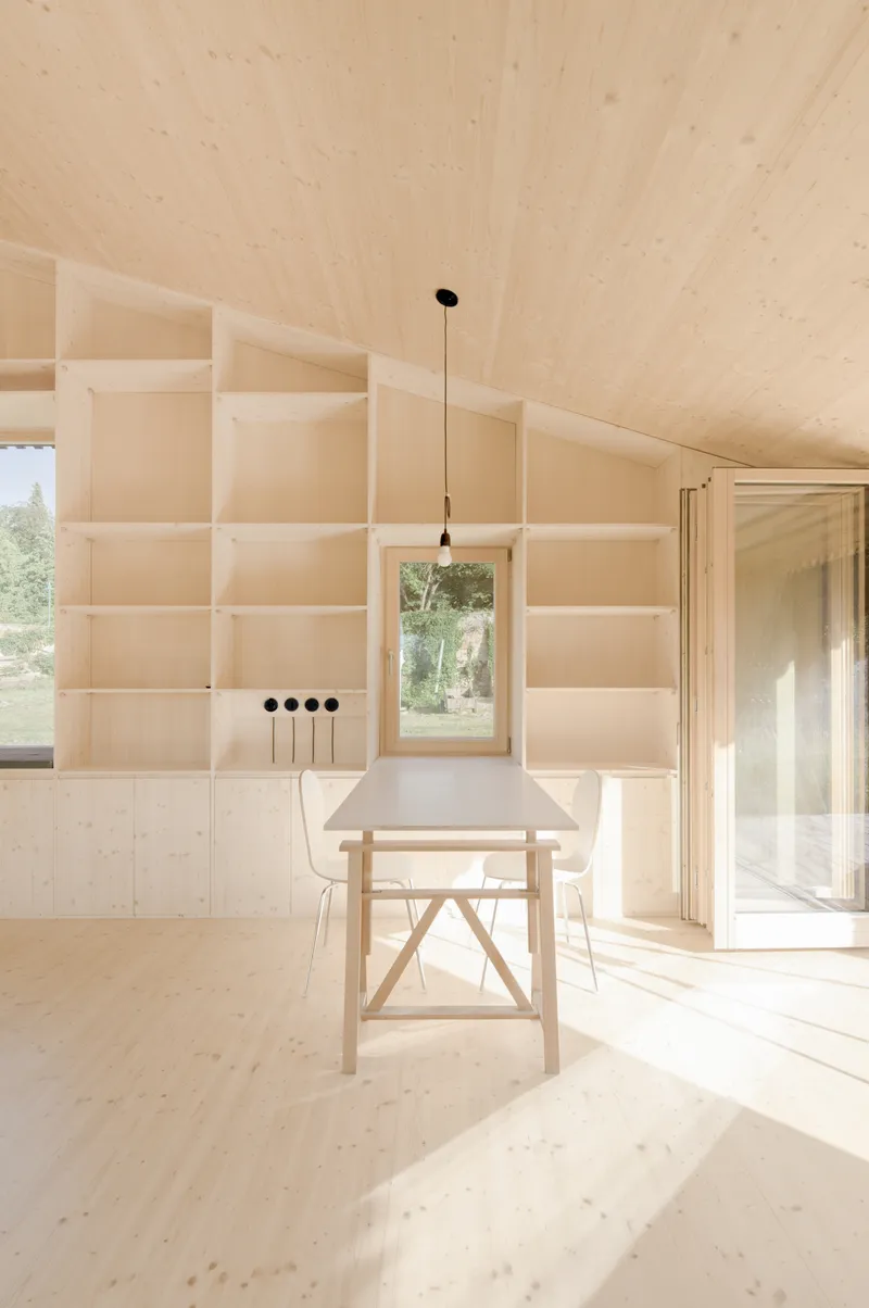 Plywood table and pendant light beneath sloped ceiling with built-in shelving and framed window