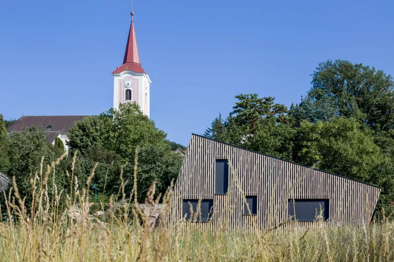 View of the timber-clad volume across the meadow with a church steeple beyond