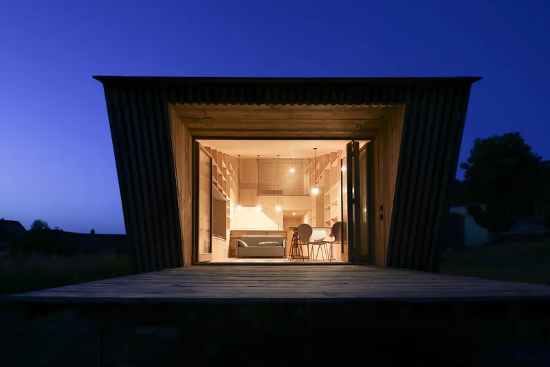 Open timber-lined living space viewed through a large glazed opening from the wooden deck at twilight