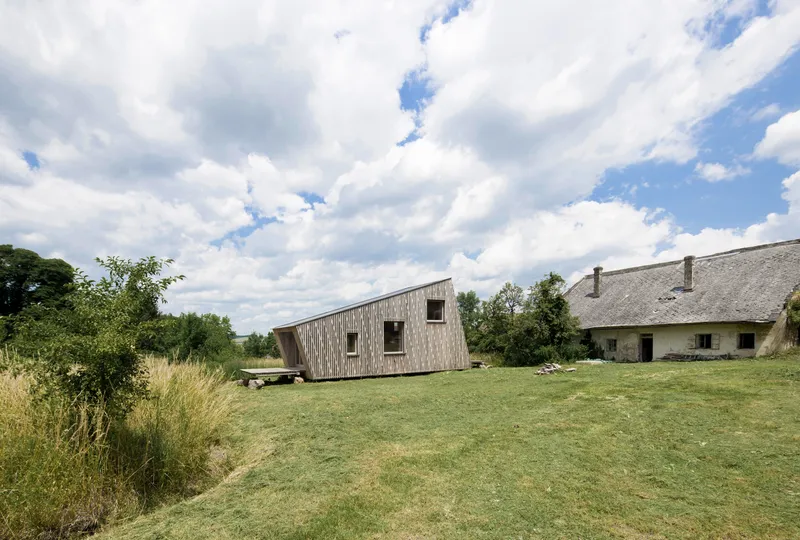 Angled timber-clad volume on a grassy lawn beside a thatched-roof building under cloudy skies