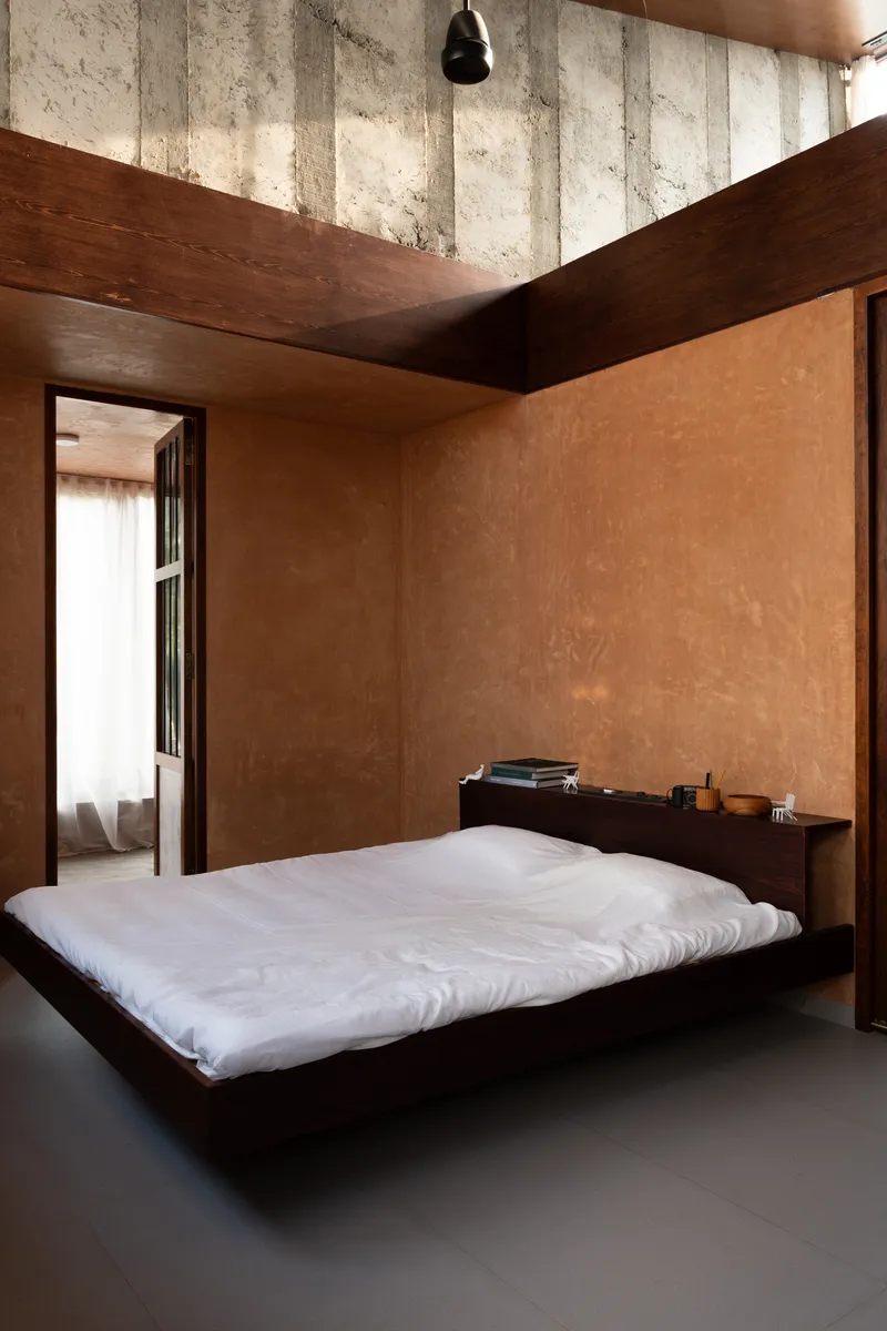 Sleeping space with low platform bed beneath exposed concrete beams and timber ceiling planes