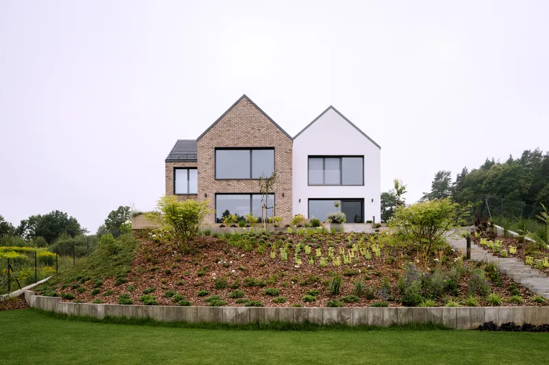Two gabled volumes with brick and white render facades above a planted hillside with concrete retaining walls