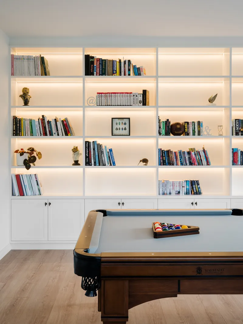 Backlit white shelving wall with books and objects above a billiard table on light wood flooring